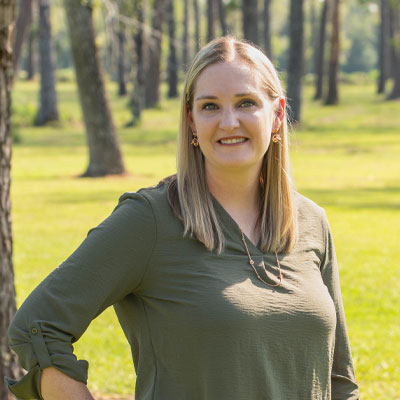 A veterinarian with shoulder-length blonde hair stands outdoors in a sunny park. She wears a green shirt, gold earrings, and smiles warmly at the camera. Tall trees and a grassy field are visible in the background, revealing her love for nature and animals alike.