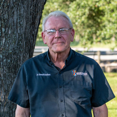 An older man with white hair, wearing glasses and a dark button-up shirt with embroidered text stands outside by a tree. As a veterinarian, he gazes at the camera with a neutral expression. The greenery and blurred background hint at his dedication to nature's well-being.