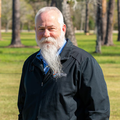 A smiling man with a long white beard and a black jacket stands in a grassy area with trees behind him. The veteran veterinarian looks towards the camera, exuding warmth and wisdom from years of dedicated service.