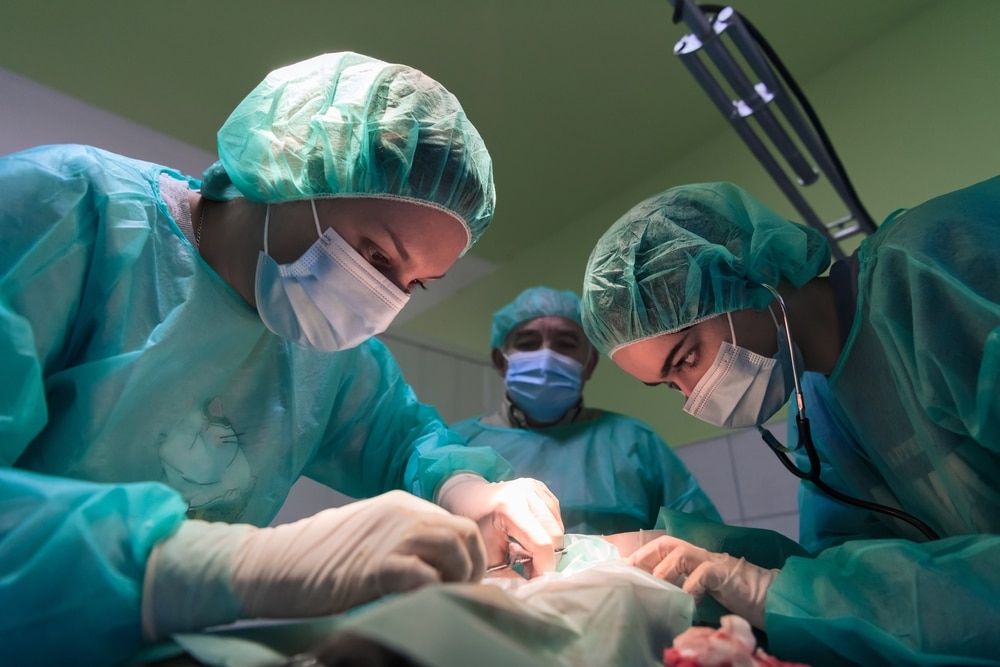 Three surgeons in green scrubs and masks perform surgery in an operating room. Two focus on the patient while one observes. Bright overhead lights illuminate the scene, emphasizing the precision required in the procedure.