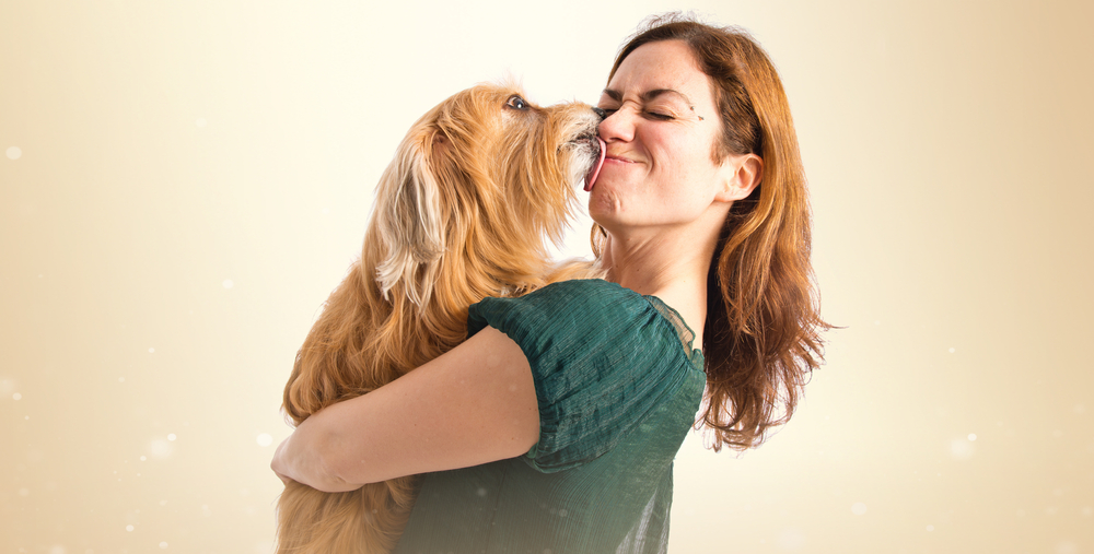 A woman in a green shirt is smiling as she holds a fluffy brown dog that playfully licks her face, reveling in the affectionate moment. The light beige background with soft, glowing bokeh effects adds warmth to this heartwarming scene, reminiscent of a caring visit to the vet.