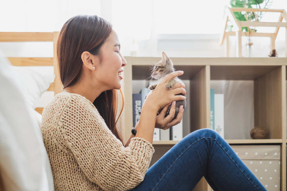 A woman, who happens to be a veterinarian, sits on the floor smiling at a small kitten she holds in her hands. She is wearing a beige sweater and jeans. Books and decor are visible in the background on a shelf. The scene is bright and cozy.