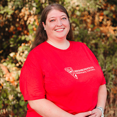 A woman with long brown hair, smiling, is wearing a red T-shirt with white text that reads "Neighborhood Veterinary Centers." She stands outdoors amid lush green foliage, embodying the friendly spirit of a dedicated veterinarian.