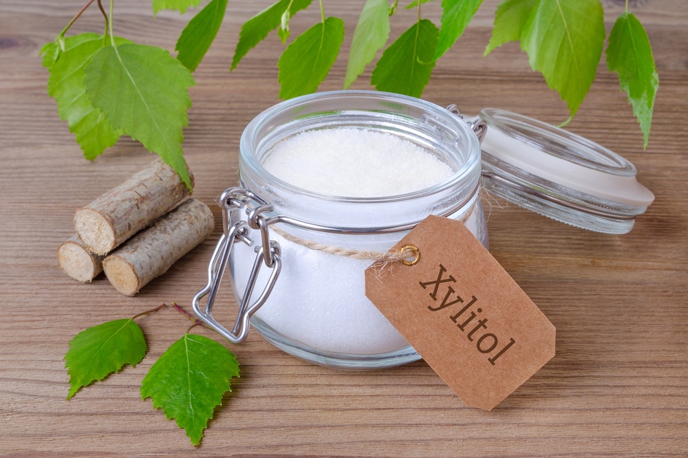 A jar of white crystalline xylitol with a labeled tag is placed on a wooden surface. Green leaves and small birch logs are nearby, suggesting a natural theme. The jar has a metal clasp and lid, and some leaves hang above.