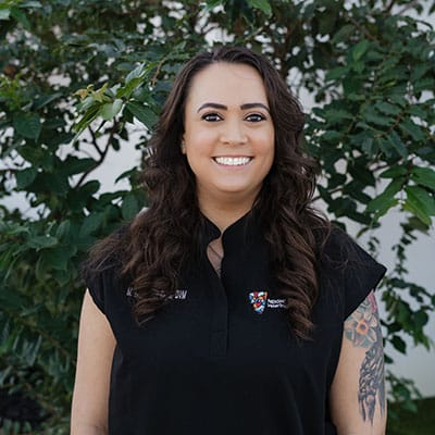 A woman with long, dark hair smiles at the camera. She is wearing a black shirt with a logo on the chest. Behind her is a backdrop of lush green leaves.